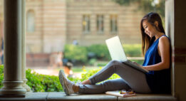 College student sitting outside on a bench with her laptop and a book and glasses by her side